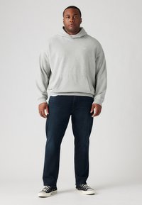 Grijze hoodie met een voorzak en een capuchon met trekkoord, gecombineerd met donkerblauwe jeans en zwarte sneakers met witte zolen.