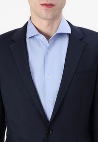 Donker marineblauwe blazer met een enkele knoopsluiting en reverskraag over een lichtblauw, overhemd met knopen. Soepele textuur en nauwsluitende pasvorm.