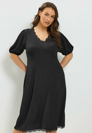 Donna con capelli lunghi che indossa un vestito nero con maniche a sbuffo e rifiniture in pizzo sullo scollo e sull'orlo, su uno sfondo chiaro e uniforme.