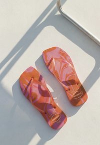 Tongs à motifs orange vif et rose sur une surface blanche avec des ombres métalliques à proximité, montrant clairement la taille et les détails de la marque.