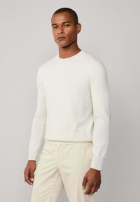 Muškarac u uskoj bijeloj dugoj majici i svijetlosmešavim hlačama, stoji naslonjen na običnu svijetlosivu pozadinu.