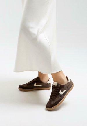 Brown and beige sneakers worn with a white satin midi skirt against a plain white background.