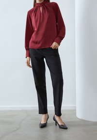 Blusa bordeaux con un'apertura a chiave e maniche lunghe, abbinata a pantaloni neri su misura e scarpe nere con tacco alto. Tessuto morbido.