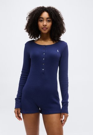 Mladá žena s kučeravými vlasmi, oblečená v tmavomodrom dlhom rukáve s gombíkmi, stojaca na bielom jednoduchom pozadí.