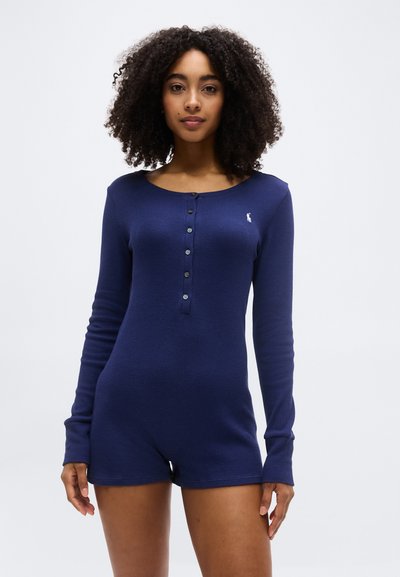 Jeune femme aux cheveux bouclés portant une combinaison bleu marine à manches longues et boutonnée, debout devant un fond blanc uni.