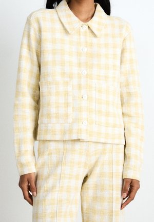 Kobieta ubrana w żółto-białą kratowaną kurtkę zapinaną na guziki z kołnierzykiem oraz pasujące spodnie, stojąca z rękami luźno opuszczonymi wzdłuż ciała.