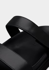 Μαύρο δερμάτινο σανδάλι με φαρδιές επίπεδες λουρίδες, ομαλή υφή και μινιμαλιστικό σχεδιασμό. Η σόλα είναι σε στερεό μαύρο χρώμα με ελαφριά καμπύλη.