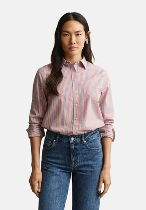 Frau mit langen dunklen Haaren, die ein pink-weiß gestreiftes Hemd mit Knöpfen und blaue Jeans trägt, steht mit entspannten Armen nach vorne gerichtet.