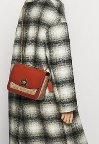 Czerwona skórzana torba crossbody z wzorzystym brązowym dnem, złotymi akcentami i kwiatowym zapięciem, noszona na tle koszuli w kratę.