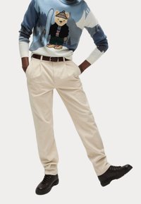 Osoba ubrana w beżowe kordowe spodnie, czarne buty na sznurowadła oraz niebieski sweter z nadrukiem misia trzymającego snowboard.