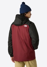 Giacca impermeabile rossa e nera con cappuccio, dotata di polsini elastici e logo sulla parte superiore della schiena. Cappello lavorato a maglia blu navy con pompon.