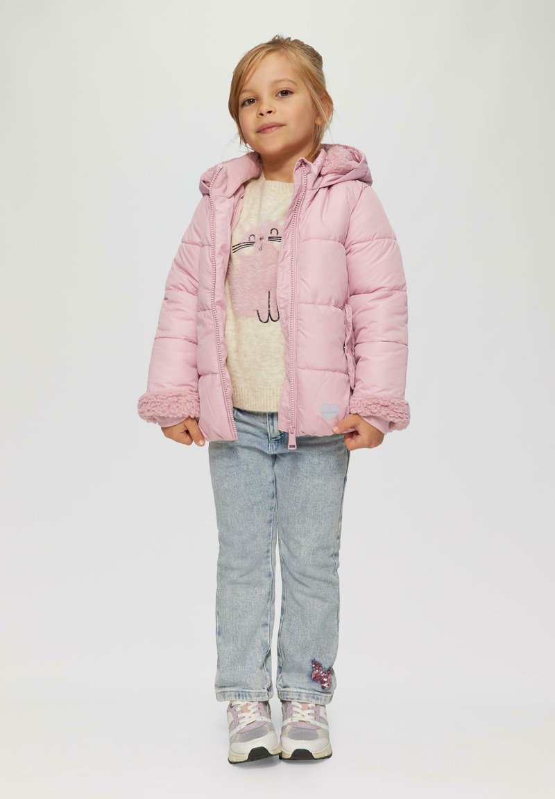Puffige rosa Jacke mit Kapuze, ausgestattet mit strukturierten Bündchen, kombiniert mit einem beigen Oberteil, das eine Katzengrafik zeigt, und hellblauen Jeans.