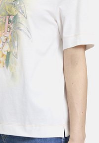 T-shirt en coton blanc à manches courtes, avec un imprimé floral coloré et des accents argentés. Fentes latérales au bas, texture douce.