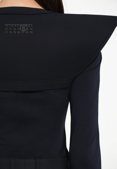 A blusa azul-marinho apresenta um colarinho estruturado e oversized, uma textura suave e uma escala de medição impressa no ombro traseiro.