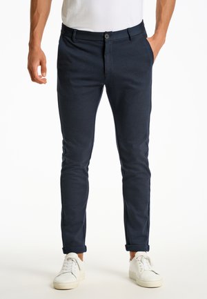 Slim-fit marineblauwe herensbroek gemaakt van zachte stof, met een knoopsluiting aan de voorkant, schuine zakken en omgeslagen boorden. Draagt witte sneakers.