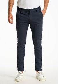 Dunkelblaue Baumwollmischung-Chinos mit schlanker Passform, ausgestattet mit einem Knopfverschluss, Seitentaschen und umgekrempelten Säumen, kombiniert mit weißen Sneakers.