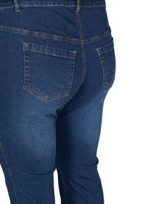 Lähikuva tummansinisten farkkujen takataskuista ja saumausten yksityiskohdista vyötäröstä reisiin henkilöllä.