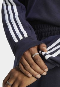 Mornarski dukserica s bijelim trakama na rukavima; rebrasti manžeti i pojas; izbliza prikazane ruke s srebrnim prstenjem, prikazujući detaljne teksture.