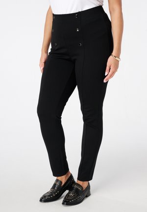 Vrouw draagt zwarte, hooggetailleerde broek met knopen en zwarte loafers met studs op een witte achtergrond.