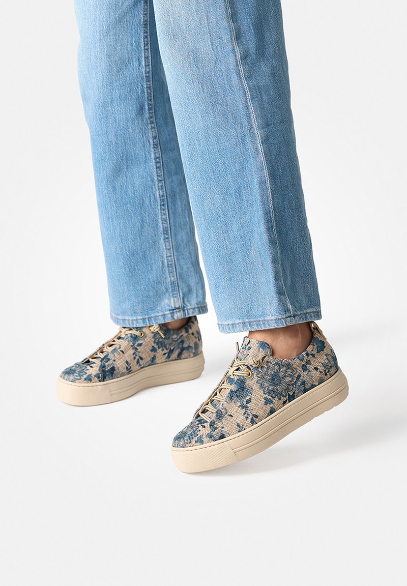 Person iført blå sneakers med blomsterprint, beige såler og lyseblå denimjeans, stående mod en ensfarvet hvid baggrund.