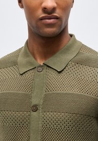 Κοντινό πλάνο ενός ατόμου που φοράει πουκάμισο πόλο από πλεκτό ελιάς με υφή, διάτρητα σχέδια και δύο καφέ κουμπιά κουμπωμένα.