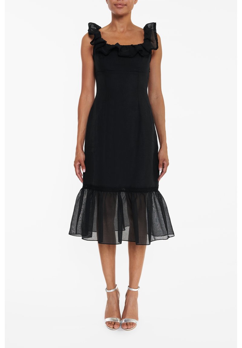 Femme portant une robe midi noire sans manches avec un col volanté et un ourlet évasé transparent, associée à des talons hauts argentés à bride cheville.