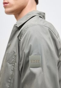 Homme portant une veste gris clair avec un patch logo carré BOSS sur la manche supérieure, col visible et poche poitrine.