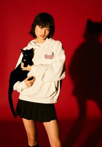 Biała bluza z kapturem z logo "Emily the Strange", czarna plisowana spódnica oraz czarny kot z żółtymi oczami na czerwonym tle. Widoczny cień.