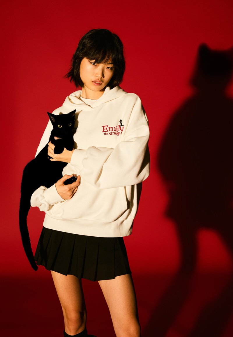 Biała bluza z kapturem z logo "Emily the Strange", czarna plisowana spódnica oraz czarny kot z żółtymi oczami na czerwonym tle. Widoczny cień.