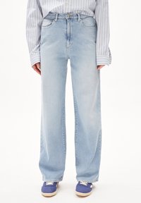 Person iført lyseblå, bredbenede jeans, stribet langærmet skjorte stoppet ind, og blå sneakers på en hvid baggrund.