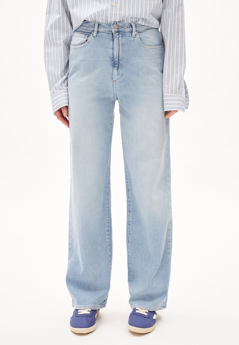 Person iført lyseblå, bredbenede jeans, stribet langærmet skjorte stoppet ind, og blå sneakers på en hvid baggrund.