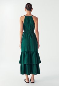 Zelena, bez rukava maxi haljina od lagane tkanine. Ima uklapajući korpus, višeslojne volane na rubu i diskretan zatvarač na leđima.