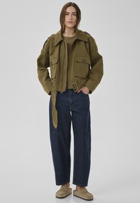 Frau trägt eine olivgrüne Jacke mit Gürtel, dunkelblaue Jeans mit weitem Bein und beigen Slippern, steht vor einem schlichten grauen Hintergrund.