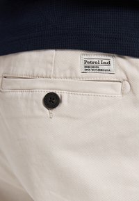 Beige bukser med en sort knap og et mærke, der står "Petrol Ind" nær en baglomme. Struktureret stof med diagonalt sømmønster.