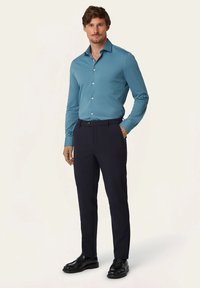 Turquoise langemouwen shirt gecombineerd met marineblauwe broek en zwarte instappers. Het shirt heeft een klassieke kraag en knopen aan de voorkant.
