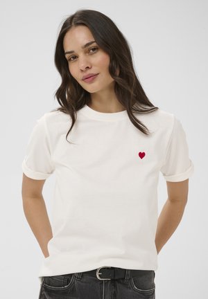 Femme aux cheveux ondulés foncés portant un t-shirt blanc avec un petit emblème de cœur rouge, debout les mains derrière le dos.