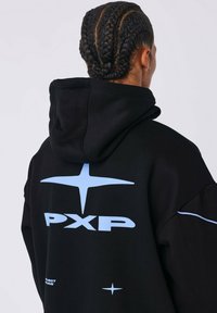 Persoon met gevlochten haar die een zwart hoodie draagt met een lichtblauwe sterlogo en de tekst "PXP" op de rug, met de rug naar de camera gekeerd.