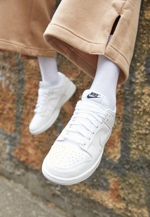 Witte leren sneakers met geperforeerde neus, platte veters en zwart Nike-logo op de tong; gecombineerd met witte sokken en beige wijde broek.