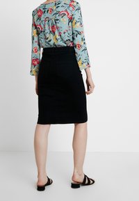 Bloemrijke blouse in lichtblauw met rode en gele patronen; zwarte spijkerrok tot de knie; zwarte sandaaltjes met bandjes; effen witte achtergrond.