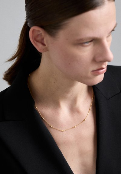 Collier en chaîne dorée avec de petites perles rondes espacées régulièrement le long de la chaîne délicate, porté sur un blazer noir. Texture lisse.