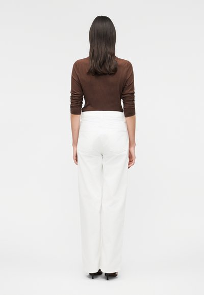 Γυναίκα με καστανά μαλλιά μέχρι τους ώμους που φοράει μία ριμπ καφέ μπλούζα με μακριά μανίκια, λευκό παντελόνι με ψηλή μέση και μαύρες γόβες, γυρισμένη με την πλάτη.