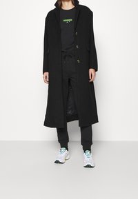Dlouhý černý kabát se zlatými knoflíky a bočními kapsami, nošený přes černý outfit, doplněný bílými teniskami s barevnými akcenty.