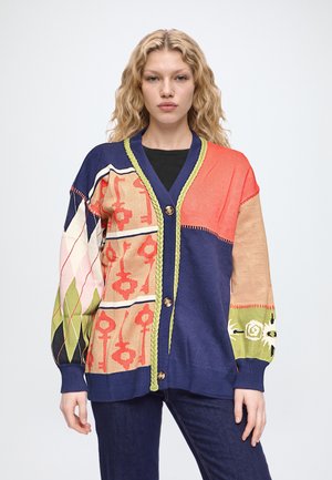 Une femme porte un cardigan patchwork coloré aux manches à motifs, avec une bordure verte et des boutons, par-dessus un haut noir et un jean foncé.