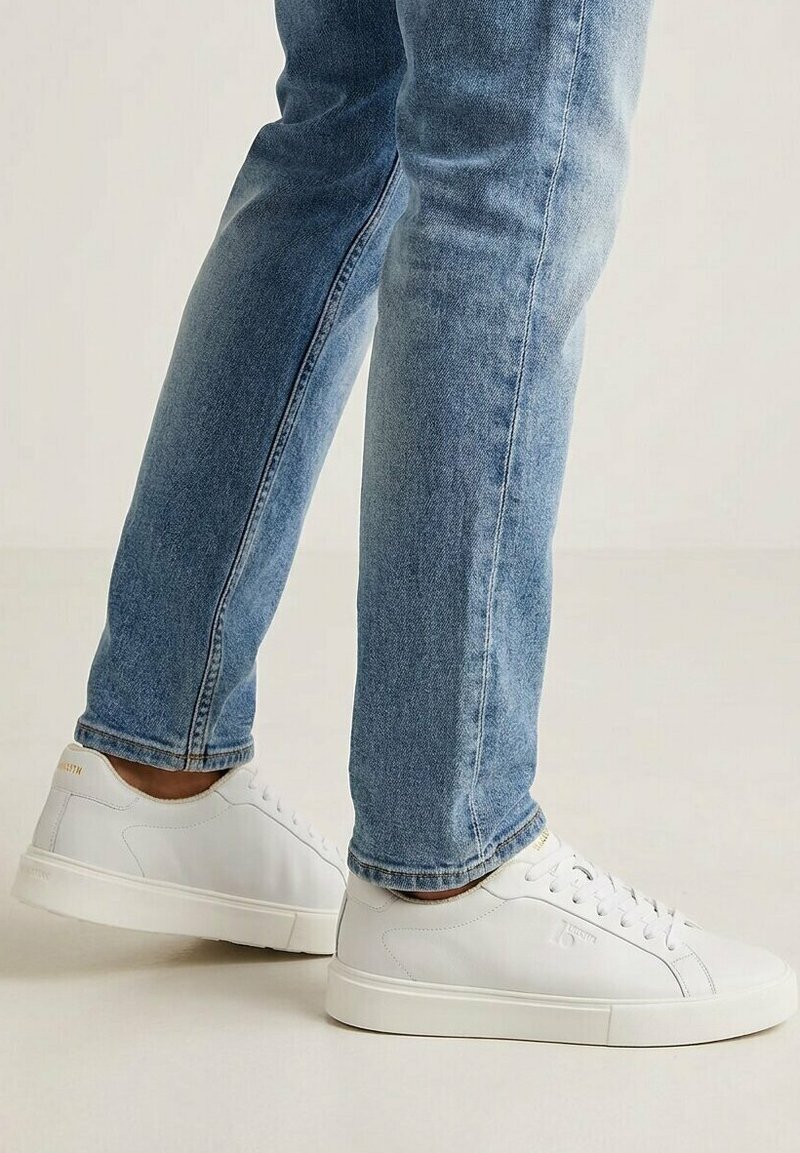 Persoon draagt lichtblauwe spijkerbroek en witte lage sneakers, staand op een lichte vloer tegen een effen achtergrond.