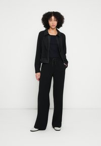 Jachetă sport neagră cu guler și șnur, asortată cu pantaloni negri cu picioare largi și un top negru mulat; textură netedă și design casual.