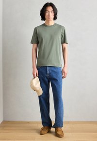 T-shirt vert olive, col rond, manches courtes ; jeans en denim bleu, coupe ample ; casquette beige à la main ; chaussures en daim marron. Fond neutre.