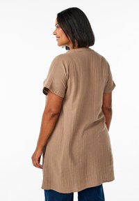 Brown, textured short-sleeve tunic with a relaxed fit, featuring a curved hem and side slits, paired with blue denim jeans.