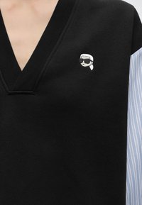 Vêtement noir à col en V avec un petit visage brodé portant des lunettes de soleil et un foulard blanc sur la poitrine, porté sur une chemise rayée bleue.