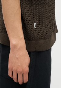 Κοντινό πλάνο αριστερής παλάμης ενός ατόμου να ακουμπάει δίπλα στο σώμα, φορώντας κορυφή πλεκτή καφέ με υφή με ετικέτα "OBEY" και μαύρο παντελόνι.