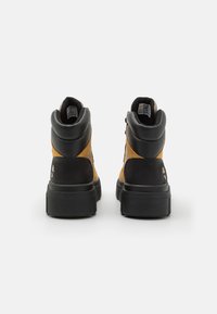 Bottes montantes avec dessus ton chameau et accents noirs, finition texturée et semelle épaisse noire. Détail de marque sur le talon.